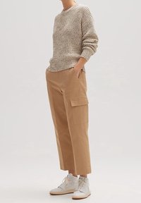 Osoba w beżowym swetrze dzianinowym, brązowych spodniach cargo z bocznymi kieszeniami i białych sneakerach typu high-top, stojąca z rękami w kieszeniach.