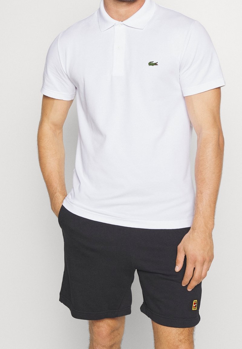 Weißes Poloshirt mit grünem Lacoste-Logo, kurzen Ärmeln und klassischem Kragen, kombiniert mit dunkelgrauen Sportshorts mit farbigem Akzent.