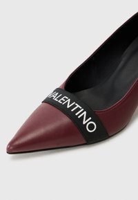 Décolleté in pelle borgogna con punta affilata, dotato di una fascetta elastica nera con la scritta bianca "VALENTINO" che attraversa la parte centrale. Texture liscia.