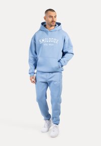 Jasnoniebieska bluza z kapturem z kieszenią z przodu i białym logo "SMILODOX ATHL WEAR". W połączeniu z pasującymi spodniami dresowymi z podobnym logo. Białe sneakersy.