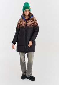 Žena oblečená v páse s potlačou leopardie a čiernej pérovke, so zelenou pletenou čiapkou, šedými džínsy a čiernymi topánkami, stojaca na pozadí z čistej bielej. 