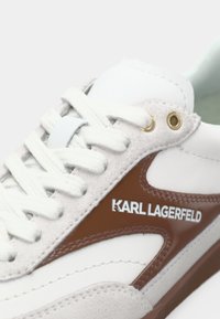 Baskets blanches avec accent en cuir marron et logo "KARL LAGERFELD", avec des lacets blancs et des œillets métalliques dorés.