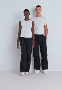 Pantaloni cargo negri cu uscătură largă, asortați cu un top alb fără mâneci și un tricou. Amândouă poartă adidași albi, stând pe un fundal neutru.