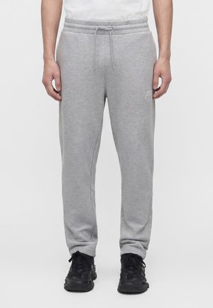 Muž v svetlosivých teplákoch so šnúrkou v páse a čiernych teniskách stojaci pred jednoduchým bielym pozadím.