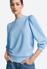 Hele sinine dressipluus puffvarrukatega, ümmarguse kaelusega ja pehme tekstuuriga. Sobitatud vabaaja teksadega, ilma nähtavate mustrite või logodeta.