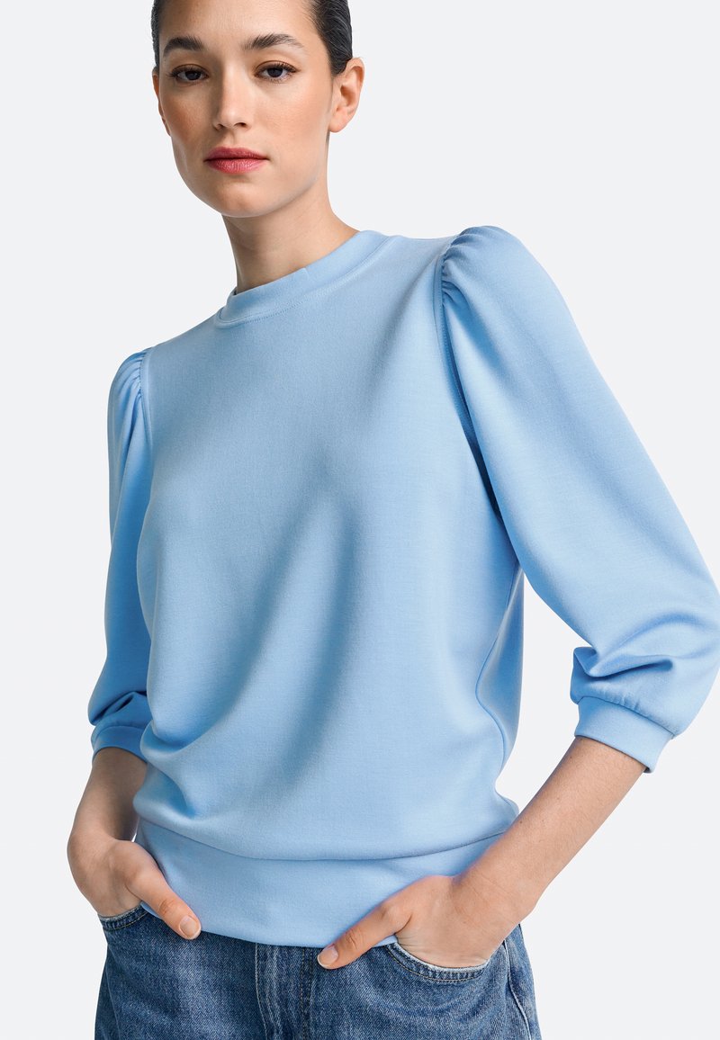 Hele sinine dressipluus puffvarrukatega, ümmarguse kaelusega ja pehme tekstuuriga. Sobitatud vabaaja teksadega, ilma nähtavate mustrite või logodeta.