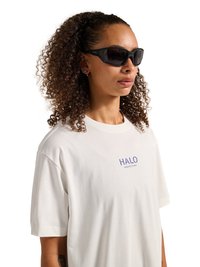 Žena sa kovrčavom kosom koja nosi crne sunčane naočale wraparound i bijelu majicu s natpisom „HALO“ na jednobijeloj pozadini.
