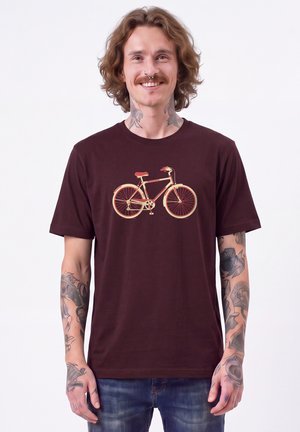 Lächelnder Mann mit Tattoos auf Armen und Hals trägt ein dunkles T-Shirt mit einer orange-gelben Fahrradgrafik und steht vor einem einfarbigen Hintergrund.