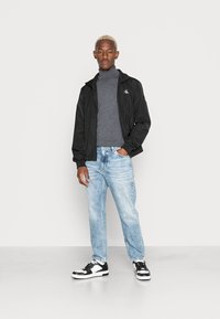 Sort letvægtsjakke over en grå polokrave, parret med lyseblå jeans og sorte og hvide sneakers. Simpel design, afslappet pasform.