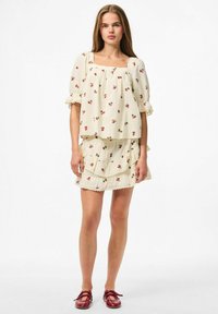 Blouse créme à manches courtes avec un motif de cerises et un décolleté carré, accompagnée d'une mini-jupe à volants. Les deux ont des bords festonnés. Chaussures rouges.