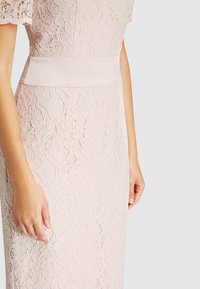 Tunikan pitsissä pehmeässä vaaleanpunaisessa, jossa on lyhyet pitsihihat, istuva vyötärö satiinivyöllä ja muotoileva siluetti kukkakuviolla.