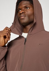 Mand iført en brun zip-up hoodie med Nike-logo, løfter huen med den ene hånd, kigger let opad og til siden.