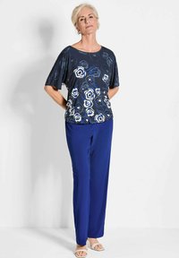 Marineblauwe bloes met korte mouwen, versierd met abstracte rozen in blauw en wit; gecombineerd met effen koninklijkblauwe broeken en lichte sandalen.