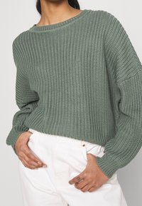 Maglione verde lavorato a maglia con una texture a coste, scollatura ampia e vestibilità rilassata. Abbinato a pantaloni di denim bianchi.