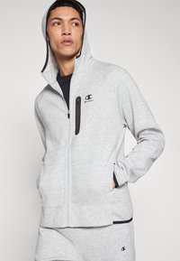 Sudadera con capucha gris con cremallera, confeccionada en tela suave, que presenta un bolsillo con cremallera negra y un logo en el pecho izquierdo. Mangas largas con puños y bajo acanalados.