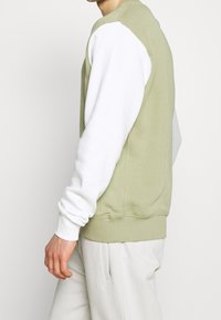 Man i ljusgrön sweatshirt med vita ärmar och ljusa byxor, stående mot en enkel vit bakgrund.