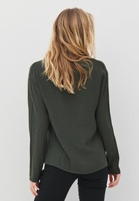 Tamsiai žalia ilgomis rankovėmis marškinėliai su laisva prigludimu, apvaliu apvadu ir minkšta tekstūra. Turintys minimalų siuvimą ir jokių matomų metalinių detalių.