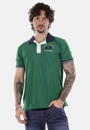 Uomo con capelli ricci e tatuaggi che indossa una polo verde con colletto blu navy e patta dei bottoni bianca, che posa con una mano in tasca.