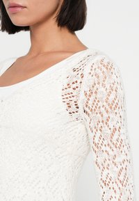 Λευκή πλεκτή μπλούζα με υφή και ανοιχτό σχέδιο, μακριά μανίκια δαντέλας και στρογγυλή λαιμόκοψη. Απαλό, ελαφρύ υλικό.