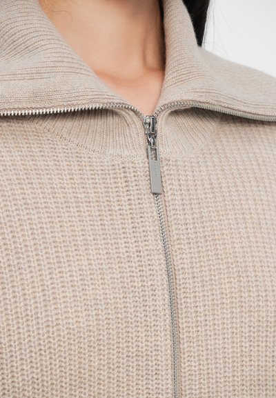 Beige hettegenser med glidelås og ribbet tekstur, samt høy krage. Har en sølvfarget glidelås og et komfortabelt strikkedesign ved halsen.