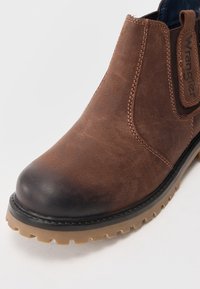 Hnědé kožené kotníkové boty s hladkou texturou, černou gumovou podrážkou a elastickými bočními panely. Na straně je viditelné logo Wrangler.