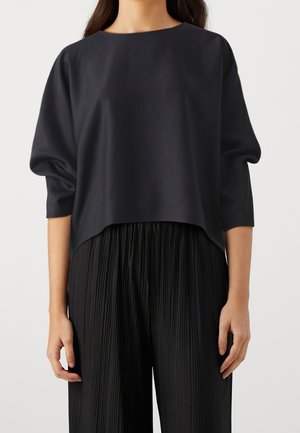 Femme portant un haut noir ample à manches longues avec un col rond et un pantalon noir plissé taille haute sur un fond uni.