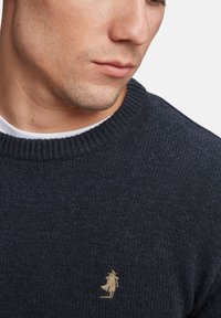 Jersey de color navy con cuello de canalé. Presenta una pequeña figura bordada en dorado en el pecho. Textura suave con un ajuste moderado.