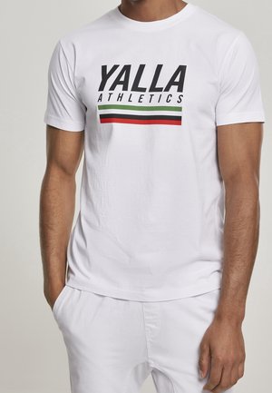 Mężczyzna ubrany w biały t-shirt z napisem "YALLA ATHLETICS" wydrukowanym na czarno nad poziomymi paskami w kolorach zielonym, czarnym i czerwonym.