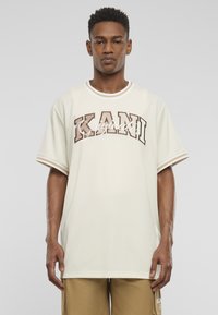 Cremefarvet oversized t-shirt med ribbet hals og brun skrift "KANI" tværs over brystet. Korte ærmer har hvide accenter.