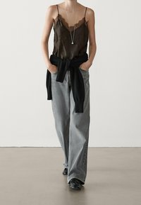 Svart spetskantad linne med tunna axelband, kombinerad med ljusgrå vida jeans. En svart tröja är knuten runt midjan. Svarta skor.