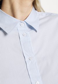 Camicia button-up a righe blu chiaro e bianche con colletto classico, dotata di bottoni bianchi e un tessuto liscio e testurizzato.
