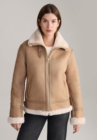TAN shearling jack met een zachte witte leren kraag, ritssluiting, zijzakken en met bont gevoerde manchet. Gladde lederen textuur over het geheel.