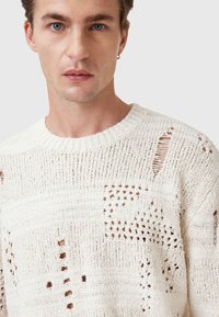 White knitted sweater with textured patterns, featuring open weaves and a square patch. Crew neck and visible stitching details.