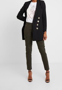 Blazer noir ajusté avec des boutons dorés, t-shirt graphique blanc et pantalon cargo vert olive, complété par des escarpins noirs à brides.