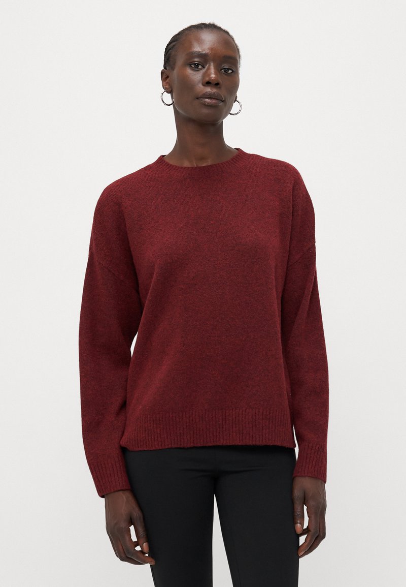 Sweater de malha vermelha com gola redonda, corte solto e detalhe canelado nas mangas e na bainha. Usado sobre calças pretas, com textura suave.