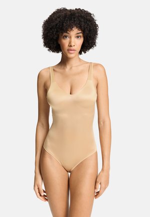 Kvinde med krøllet hår iført en glat, beige, ærmeløs bodysuit, stående mod en ensfarvet lys baggrund.