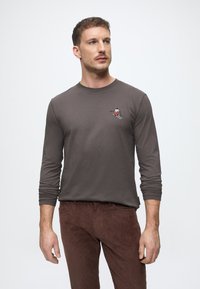 Rudas ilgomis rankovėmis marškiniai iš minkštos medžiagos su mažu siuvinėtu slidininko figura ant krūtinės, dėvimi su tamsiai rudais štrampais.