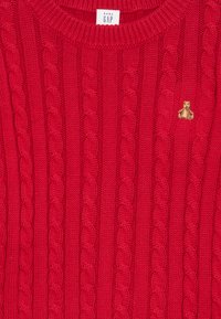 Pull en tricot rouge avec un design en câble et un col côtelé. Présente un petit accent brodé en forme d'ours en peluche au-dessus de la poitrine gauche.