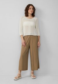 Gebreide crèmekleurige trui met ronde halslijn en driekwart mouwen, gecombineerd met tan, loszittende, wijde broek en beige sandalen met bandjes.