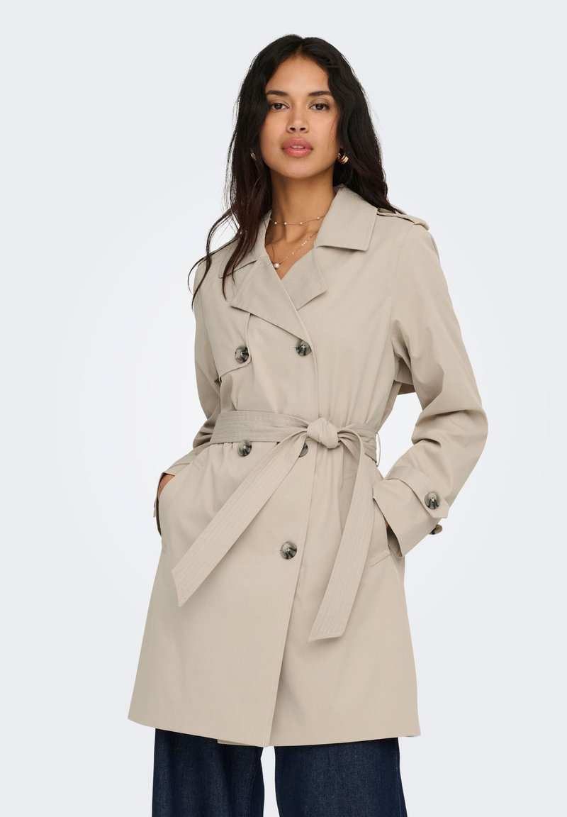 Femme portant un trench-coat beige croisé avec ceinture à la taille et poches, associé à un jean bleu foncé, debout devant un fond uni.