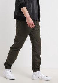 Olivengrønne, tilspidsede bukser med elastik i taljen, der har sidelommer og en glat tekstur, kombineret med hvide højtop-sneakers.