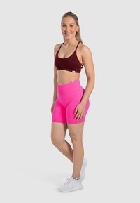 Sujetador de color rojo oscuro y pantalones cortos de talle alto en rosa brillante. La tela parece elástica; la modelo lleva zapatillas blancas. Diseño sencillo con acentos mínimos.