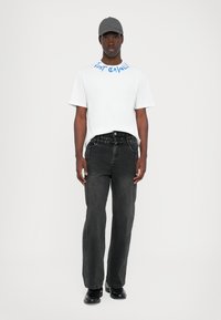Hvit t-skjorte med blå grafisk tekst, kombinert med svarte vide jeans med sidesøm. Fullført med en grå caps og svarte sko.