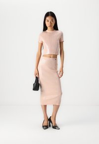 Roze sjajni crop top i odgovarajuća midi suknja s rebrastom teksturom. Model drži malu crnu torbicu i nosi crne štikle s metalnim naglascima.