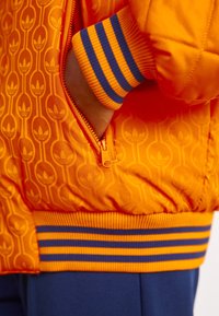 Orange quilted bomber jacket with a repeating logo pattern, featuring a zippered pocket and ribbed cuffs in blue and orange stripes.