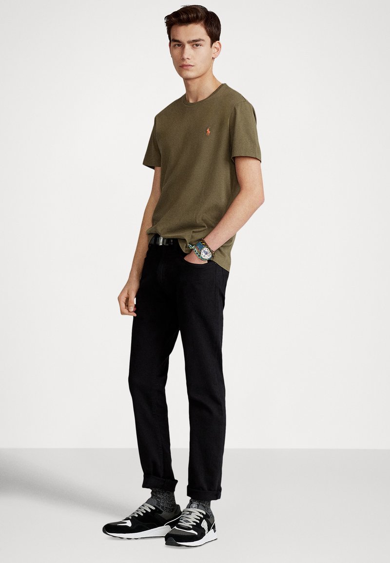 Olive green short-sleeve t-shirt with a small emblem, black trousers, and black sneakers with gray accents, showcasing a casual look.