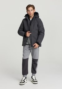 Czarna ocieplana kurtka z kapturem, noszona na szarej bluzie i dopasowanych spodniach dresowych z szarymi i ciemnoszarymi blokami kolorów. Czarne sneakersy.