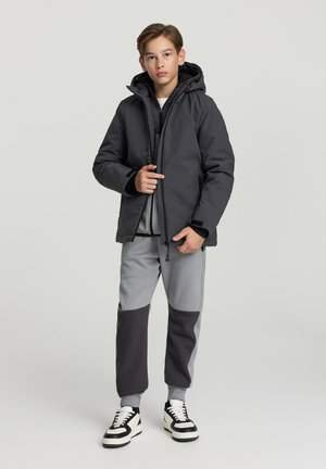 Giacca nera imbottita con cappuccio, indossata sopra una felpa grigia e pantaloni della tuta abbinati con blocchi di colore grigio e grigio scuro. Sneakers nere.