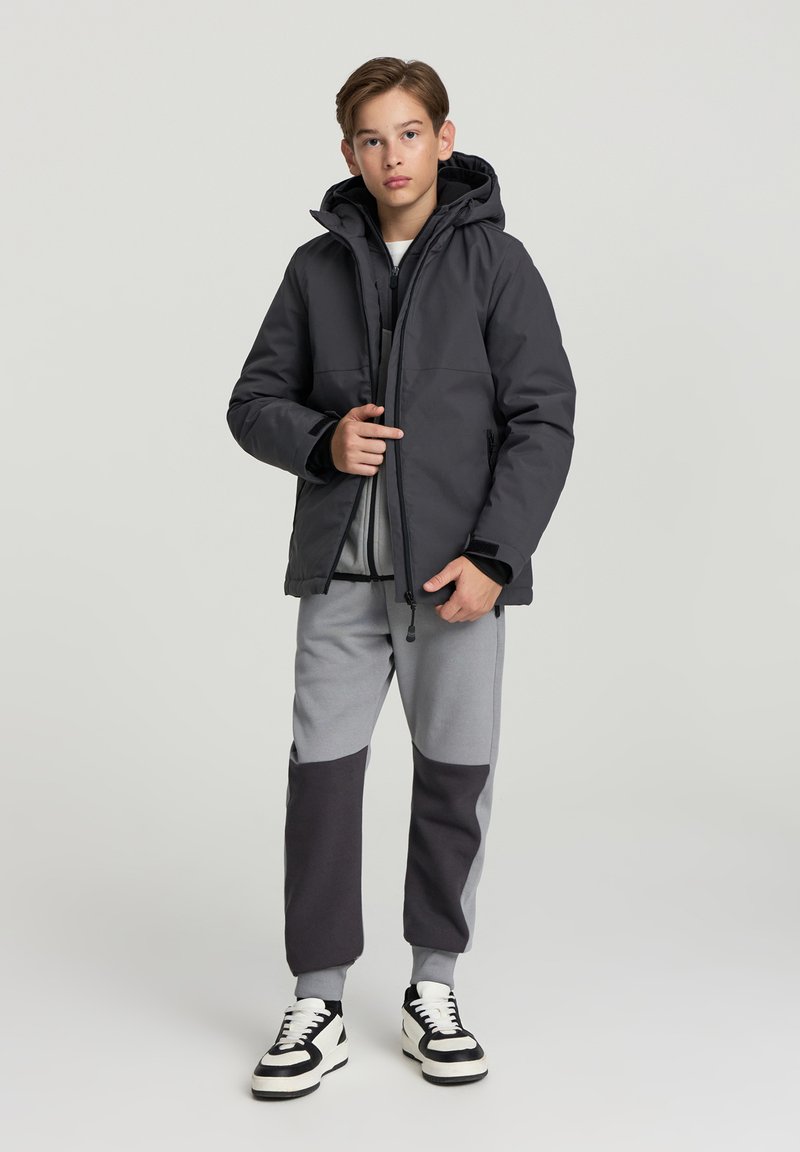 Czarna ocieplana kurtka z kapturem, noszona na szarej bluzie i dopasowanych spodniach dresowych z szarymi i ciemnoszarymi blokami kolorów. Czarne sneakersy.