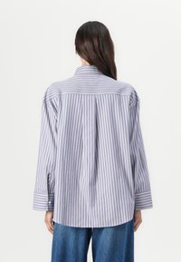 Camicia a maniche lunghe a righe verticali in lilla chiaro e bianco, con piega sul retro e polsini con bottoni. Il tessuto sembra leggero.